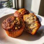 Three delicious banana nut muffins on a white plate, one cut in half showing the moist interior texture.