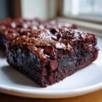 A close-up of a rich, fudgy slice of black bean brownies topped with melted chocolate chips on a white plate.