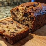 A freshly baked, moist date nut bread loaf with one slice cut and resting beside it on a wooden board.