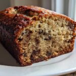 Close-up of a moist slice of greek yogurt banana bread with visible chocolate chips on a white plate.
