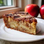 A close-up of a moist slice of jewish apple cake topped with cinnamon sugar, served on a white plate with red apples in the background.