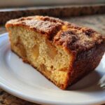 A close-up of a moist slice of jewish apple cake with visible apple chunks, served on a white plate.