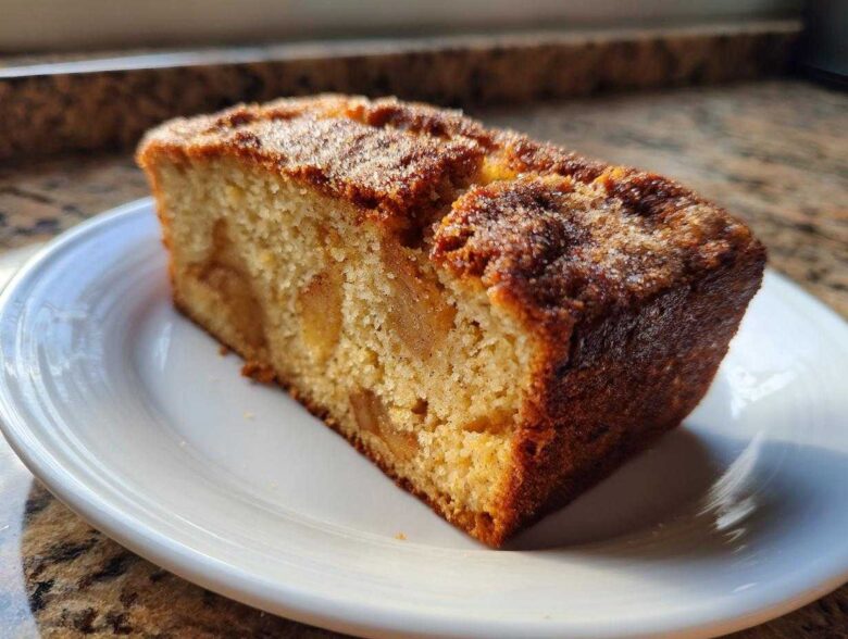 A close-up of a moist slice of jewish apple cake with visible apple chunks, served on a white plate.
