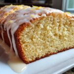 Close-up of a moist lemon loaf, sliced to show the crumb, generously covered in dripping lemon glaze and zest.