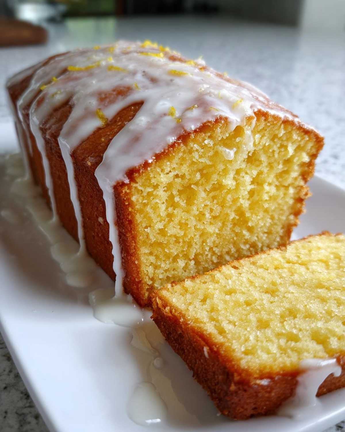 A close-up of a moist lemon pound cake loaf, partially sliced, topped with a thick white glaze and lemon zest.