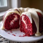 A slice cut from a moist red velvet bundt cake showing a cream cheese swirl center, topped with white glaze.