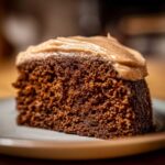 Close-up of a moist slice of dark gingerbread cake topped with thick, creamy brown frosting.