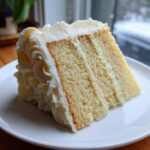 Close-up of a moist, three-layer slice of vanilla cake recipe covered in thick white buttercream frosting.