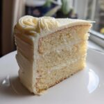 A perfectly moist slice of vanilla cake recipe with thick white frosting, sitting on a white plate.