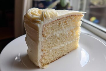 A perfectly moist slice of vanilla cake recipe with thick white frosting, sitting on a white plate.
