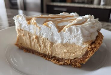 A thick slice of no-bake peanut butter pie showing graham cracker crust, creamy filling, and whipped topping drizzled with peanut butter.