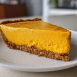 A thick, bright orange slice of no bake pumpkin pie with a dark graham cracker crust served on a white plate.