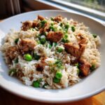 Close-up of a white bowl filled with savory one pot chicken and rice mixed with bright green peas and herbs.