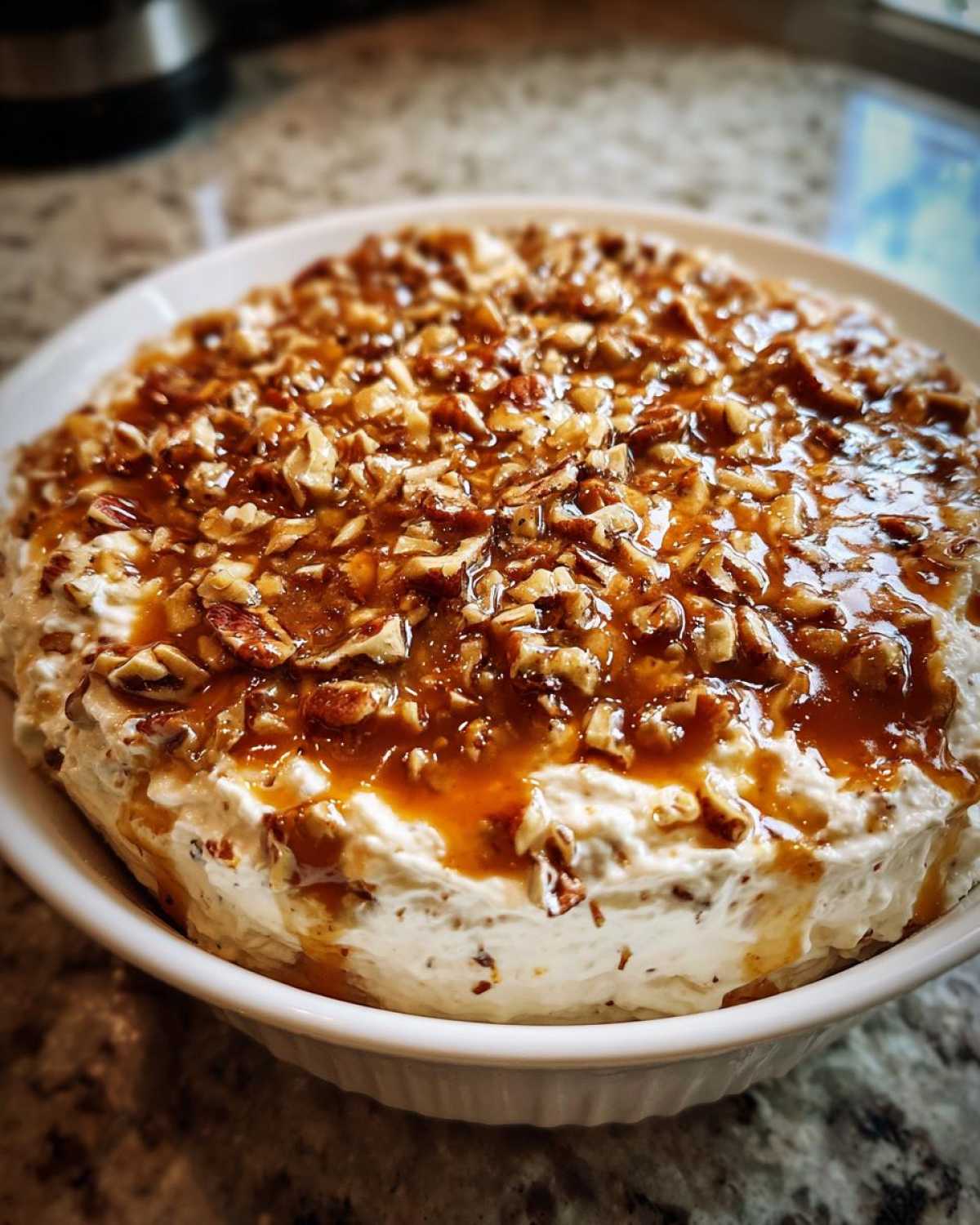 A close-up of a creamy pecan pie dip in a white bowl, generously topped with chopped pecans and thick caramel sauce.