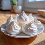 A close-up of several perfectly piped, crisp white meringue cookies stacked on a small white plate.