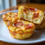 Close-up of golden-brown pizza muffins topped with melted cheese and pepperoni, one muffin is cut open.