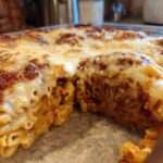 Close-up of a serving removed from a baked pizza pasta bake, showing melted cheese and tubular pasta.