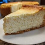 A thick, creamy slice of baked Protein Cheesecake with a golden top and graham cracker crust, served on a white plate.