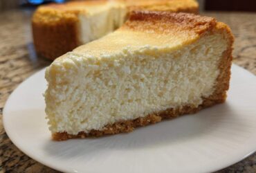 A thick, creamy slice of baked Protein Cheesecake with a golden top and graham cracker crust, served on a white plate.