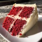A moist slice of vibrant red velvet cake layered and covered in thick white cream cheese frosting.