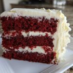 A close-up of a thick slice of red velvet cake with three layers and cream cheese frosting.