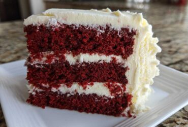 A close-up of a thick slice of red velvet cake with three layers and cream cheese frosting.
