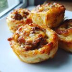 Close-up of four golden brown sausage pinwheels topped with melted, bubbly cheese on a white plate.