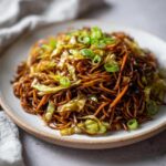A close-up of a plate piled high with dark, savory fried noodles mixed with cabbage and topped with sliced green onions.