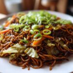 Close-up of a plate piled high with dark, savory fried noodles mixed with cabbage and carrots, topped with fresh green onions.