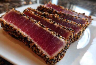 Four slices of perfectly cooked Seared Ahi Tuna, crusted with black and white sesame seeds, resting on a white plate.