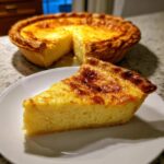 A slice of rich, golden baked egg custard pie served on a white plate with the rest of the pie in the background.