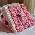 A tall slice of pink layer cherry cake with white frosting and visible whole cherries on a white plate.
