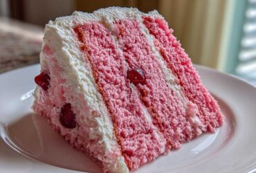 A tall slice of pink layer cherry cake with white frosting and visible whole cherries on a white plate.