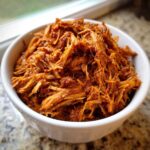 Close-up of shredded, saucy Slow Cooker Pulled Pork piled high in a white bowl.