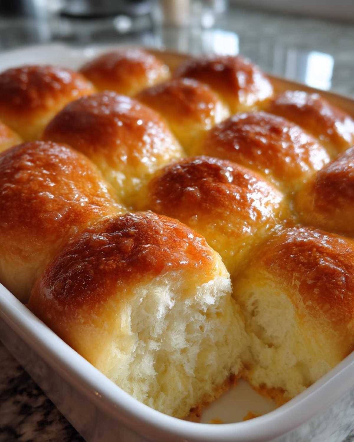 A close-up view of freshly baked, golden-brown dinner rolls, glistening with a buttery glaze, one roll pulled apart showing the soft interior.