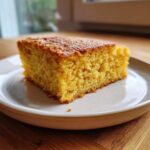 A close-up of a moist, golden slice of sourdough cornbread served on a light-colored plate.