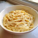 A white bowl filled with spaghetti generously coated in creamy Olive Garden Alfredo Sauce, topped with pepper.