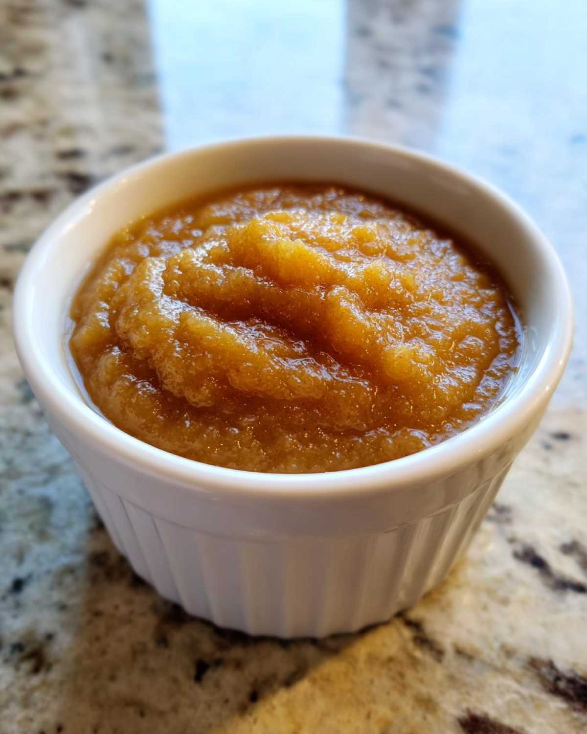 Close-up of rich, textured homemade applesauce served in a small white ramekin on a granite counter.
