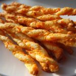 A close-up of a stack of golden, twisted, spicy Southern cheese straws resting on a white plate.