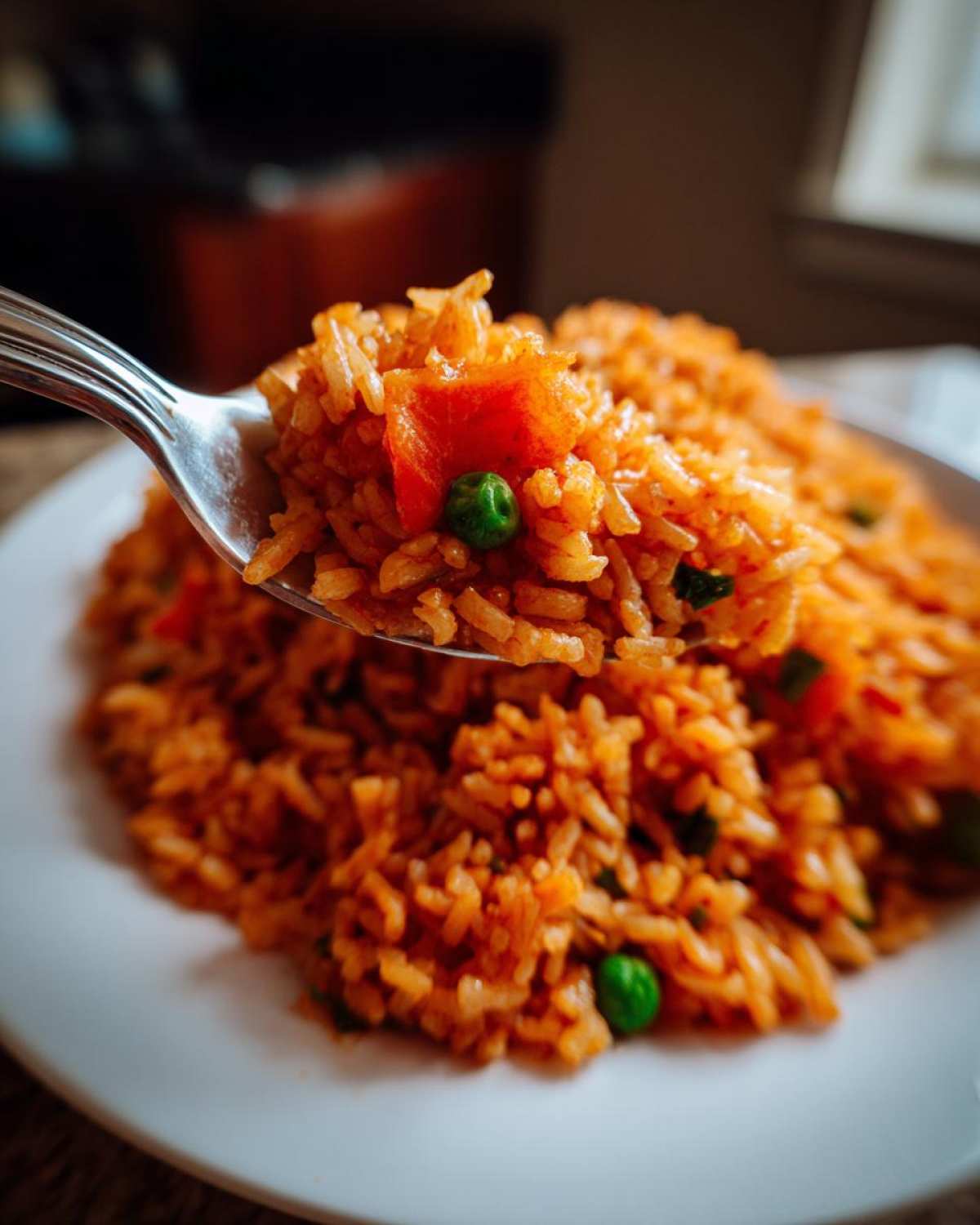 A spoonful of bright orange Spanish rice, featuring peas and diced tomato, lifted above a plate of the dish.