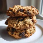 A close-up stack of three golden brown oatmeal raisin cookies resting on a white plate.