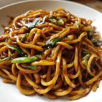 A close-up view of thick, glossy, brown sticky garlic noodles garnished with fresh green scallions.