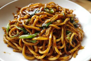 A close-up view of thick, glossy, brown sticky garlic noodles garnished with fresh green scallions.