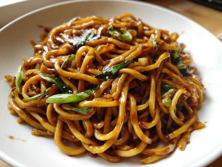 A close-up view of thick, glossy, brown sticky garlic noodles garnished with fresh green scallions.