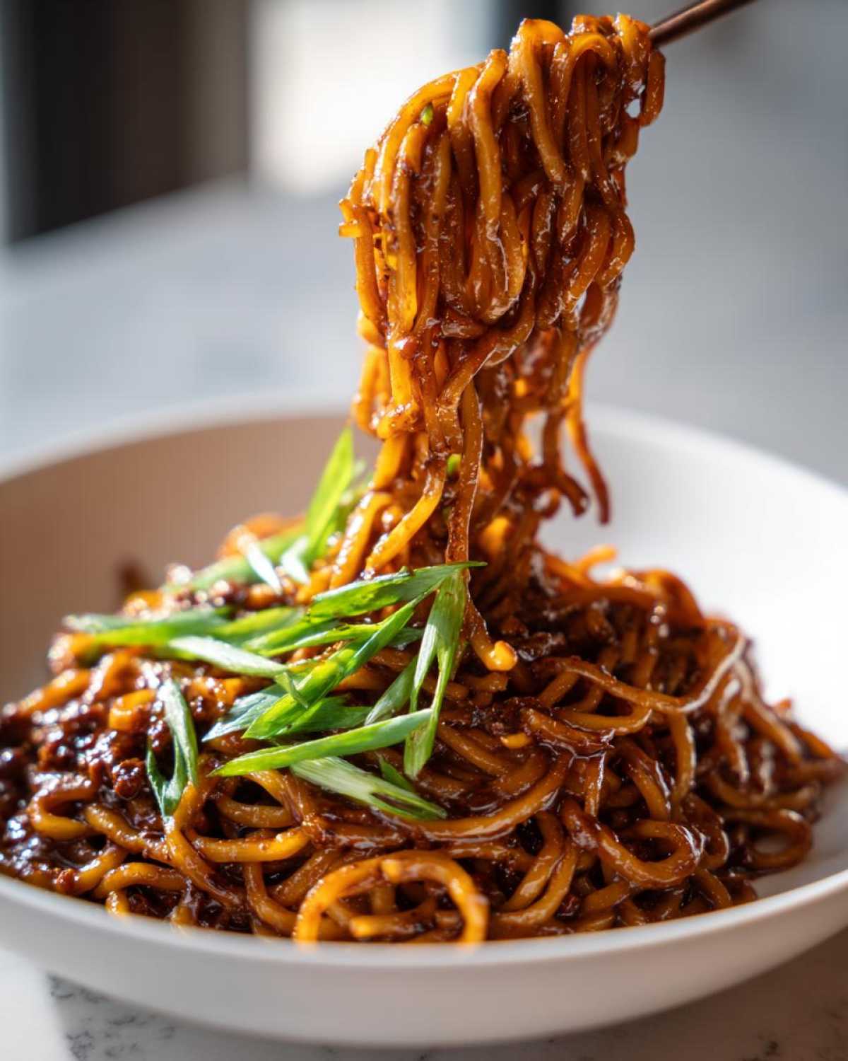 Close-up of sticky garlic noodles being lifted high from a white bowl with chopsticks, topped with fresh green scallions.