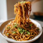 A close-up shot of sticky garlic noodles being lifted high with chopsticks from a white plate, garnished with green onions.