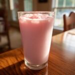 A tall glass filled with pink strawberry milk and ice cubes sitting on a shiny wooden table.