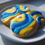 Four round sugar cookies decorated with piped royal sugar cookie icing in swirling patterns of bright blue, yellow, and white, covered in sparkling sanding sugar.