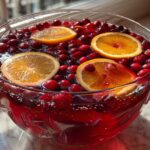 Close-up of a large glass bowl filled with red thanksgiving punch, garnished with floating cranberries and bright orange slices.
