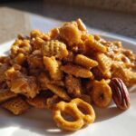 A mound of glossy, caramel-coated toffee chex mix recipe featuring cereal pieces, pretzels, and pecans on a white plate.